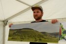 Ein Mann mit Schildkappe hält ein Panoramafoto vor sich. Dieses zeigt einen querterrassierten Weinberg.