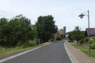 Ein gepflasterter Gehsteig flankiert eine neu geteerte Straße. Sie führt zur Ortsmitte, wo der Turm einer modernen Kirche aufragt.