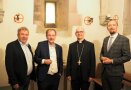 ALE-Leitung und ihr bischöflicher Besuch reihen sich zu viert zum Gruppenbild in einer historischen Kapelle auf. Aus der Wand ragen drei schmiedeeiserne Kerzenhalter.