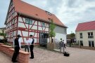 Vor dem restaurierten Fachwerkhaus befinden sich vier Parkplätze und ein junger Obstbaum. Drei Männer, zwei davon in traditionelle Zimmermannskluft, unterhalten sich.