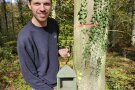 Ein junger Mann in Arbeitskleidung steht neben einem mit Efeu berankten Bau und hält einen Vogelnistkasten, der vorne offen ist.