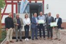 Sieben Menschen stehen vor dem Eingang des Hauses. Die einzige Frau hält einen großen gebastelten Schlüssel in der Hand. Rechts von ihr präsentieren jeweils zwei Männer grüne Projekt- und Fördertafeln.