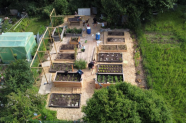 Ein Nutzgarten aus der Vogelperspektive. Er besteht aus normalen und einigen Hochbeeten – insgesamt 14 Stück sowie einem Gewächshaus. Der begehbare Bereich ist mit Hackschnitzeln bedeckt.