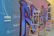 Ein Mann auf einer hölzernen Stehleiter sprüht mit der Farbe Blau das Wort RECHTE an eine Wand.