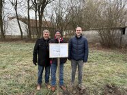 Drie Personen stehen an einem Schild. Hier entsteht ein Regenrückhaltebecken.