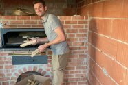 Ein junger Mann hat die Hände voller dünner Holzscheite. Er steht an einem mit Ziegelsteinen frisch gemauerten Backofen.