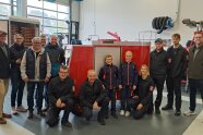 Im Feuerwehrgerätehaus steht ein rotes Einsatzfahrzeug, das geschrumpft wirkt. Mädchen und Jungen, Frauen und Männer mit und ohne Uniform umringen das Kinderfeuerwehrauto.