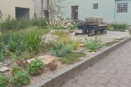 Einladend wirkt eine mit Sträuchern, Blumen, Gräsern und Kräutern bepflanzte Fläche. Dazwischen steht eine Lore auf einem kurzen Schienenstück. Sie ist mit Steinquadern beladen.