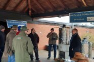 Eine mobile Brauanlage steht im Vordergrund, umgeben von Menschen, die unter fachkundiger Anleitung gemeinsam Bier brauen. 