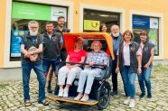 Die Chauffeure der Seniorenrikscha tragen eine Weste mit dem Logo der Nachbarschaftshilfe Gerolzhofen. Vor deren Laden haben sie sich zusammen mit zwei Fahrgästen aufgestellt.