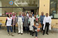 Ein gutes Dutzend ziemlich schräg wirkender Menschen in Schürzen, Latzhosen und weißen Arbeitsmänteln haben sich vor einem Schaufenster postiert. Über ihnen ist der dreizeilige Schriftzug „else! | Waschsalon | PopUp Galerie“ zu lesen.