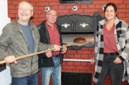 Eine Frau und zwei Männer stehen mit einem Brotschieber an der offenen Tür eines gemauerten Backofens.