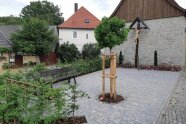 Ein Platz von der Größe etwa der Grundfläche eines Einfamilienhauses ist mit glatten Pflastersteinen belegt. Er wird auf der Stirnseite vom Giebel einer Scheune begrenzt. Gegenüber und auf einer Längsseite befinden sich Blumen- und Sträucherrabatten mit Parkbänken davor. Auf der zweiten Längsseite schließt ein helles Kopfsteinpflaster an. In der Mitte ist in eine Baumscheibe eine junge Platane in vollem Grün eingepflanzt. Vor der Scheunenwand steht ein hohes Kreuz aus dunklem Holz mit hellem Corpus.