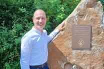 Ein Mann mit schütterem Haar greift an einen Felsbrocken. Daran ist eine Bronzeplatte befestigt, die auf die Flurneuordnung und Dorferneuerung in Oberweißenbrunn verweist.