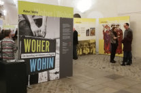 Eine Person hält einen alten Koffer mit beiden Händen. Darauf stehen groß die Worte „WOHER“ und „WOHIN“, getrennt durch einen weißen Balken mit der Schrift „Eine Ausstellung vom Ankommen und Weggehen“