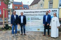 Zwei Vertreter des ALE Unterfranken sowie der Auraer Bürgermeister und die Gottesdienstleiterin der katholischen Kirche stehen vor einem Bauzaun, an dem ein Transparent mit den offiziellen Angaben zur Dorferneuerung aufgespannt ist.