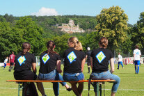 Eine Personengruppe sitzt am Spielfeldrand. Im Hintergrund ist die Karlsburg zu erkennen.