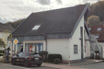 Ein eingeschossiges, helles Gebäude mit steilem, dunklem Ziegeldach steht an der Durchgangsstraße. Es ist barrierefrei zugänglich.