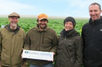 Vier Personen vor einem grünen Feld werben mit einem Schild die staatliche Initiative „boden:ständig“