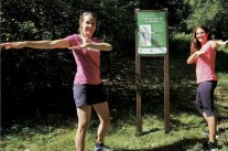 Zwei Frauen in Sportkleidung machen Schulterdehnungsübungen. Zwischen ihnen ist ein Schild an einem Pfahl angebracht. Es verweist auf den Atemlehrpfad. Ihn anzulegen, hat die Kreuzbergallianz gefördert