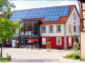 Der Dorfladen mit Mehrgenerationenwerkstatt in Aidhausen. Vor dem Gebäude stehen Stühle, Tische und ein Sonnenschirm. Das Dach des Gebäudes ist mit Photovoltaiksensoren belegt. Links und rechts im Bild steht je ein Baum. Die Fassade des Mehrgenerationenhauses ist im Erdgeschossbereich rot, im Obergeschoss weiß angestrichen.
