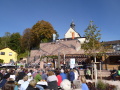 Die Erasmuskapelle mit schiefergedecktem Glockentürmchen steht erhöht über dem Dorfplatz. Der weiße Giebel mit Kreuz zeigt in Richtung Dorfplatz. Eine Treppenanlage, die sich in halber Höhe teilt und vor der Kapelle wieder zusammenläuft, führt hinauf. Der Platz ist voller Menschen.