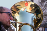 Mann spielt auf einer Basstuba: Am Wulst des Schalbechers der Tuba spiegelt sich eine Hauszeile.