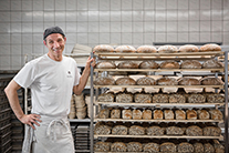 Bäcker steht neben einem mit verschiedenen Brotsorten beladenen Rollregal.