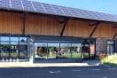 Lagerhausgebäude mit dem Dorfladen, der sich durch einen verglasten Vorbau aus Metall aus der Mauerflucht hervorhebt. Auf dem Dach ist eine Photovoltaikanlage installiert.