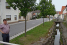 Straße im Dorf, zu der ein wasserführender, renaturierter Dorfgraben parallel verläuft. Der Straßenraum ist links und rechts bebaut. Über den Graben führen kleine Brücken zu den Häusern.