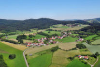 Landschaftsaufnahme aus der Vogelperspektive. Wiesen, Felder, Wälder und einige Ortschaften. Am Horizont erstrecken sich bewaldete Höhenzüge.