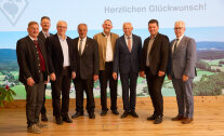 Acht festlich gekleidete Männer stehen in einer Reihe auf der Bühne