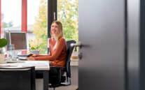 Eine junge Frau sitzt am Schreibtisch und lächelt einem entgegen
